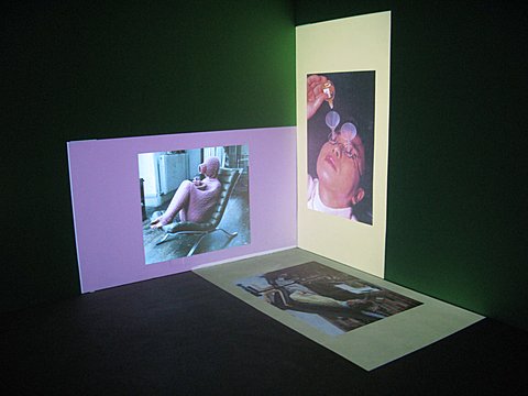 There are three images projected in the corner of a green walled room. On the floor there is a rectangular image projected with its long side against the right wall and short side against the left wall. The image is of a person and has a light yellow boarder on all sides. The image on the left wall is placed horizontally. The image itself is a square but has a boarder that makes the projection rectangular. This image is of a person in a purple-pinkish full body knitted suit sitting on a chair. The boarder of this image matches the purple-pink of the knitted garment. The image on the right wall is placed vertically. Both the image and its boarder are rectangular. The image is of a man with funnels through his glasses dropping eyedrops down the funnels. The boarder on this image is a light yellow that is more opaque than the boarder on the floor image. 