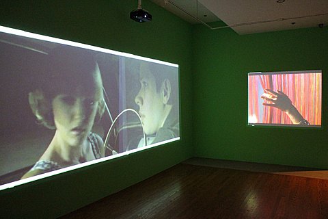 There is a corner of a green walled room with two images projected in it. On the left wall there is a large rectangular image of a man and woman in the car. On the right wall is a much smaller rectangular image of a hand brushing against colorful pink and red fabric. 
