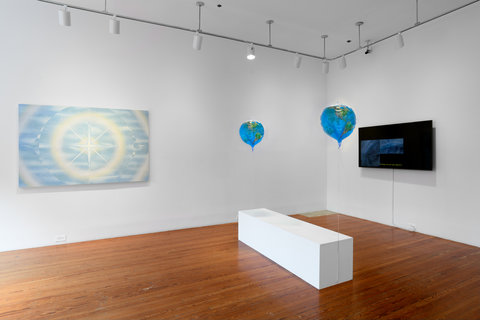 Installation image of the exhibition featuring two walls. On the left wall, a painting of blue and yellow concentric circles. A white bench sits on the floor in front of a mounted TV monitor. On either side of the bench two blue balloons float above the ground. 
