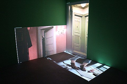 There is a corner of a green walled room with three images projected on it. The floor image is projected with its long side against the right wall and short side against the left wall. The image is of someones hands wearing gloves on a table with paperwork around them. The image on the left well is projected horizontally and shows a door in a pink walled room. The image on the right wall is projected vertically and shows two green doors that meet on their inner corners. 