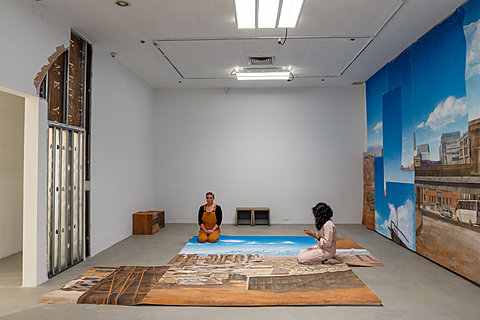 Two people kneel on a large carpet printed in color with a fragment of a landscape painting.