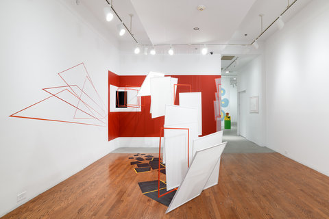 Installation view of the exhibition featuring an installation by one of the artists. On the back wall, there is a large painted red wall. The rest of the walls are painted white. The floor is made of wood. Several white sheets of paper hang from the room at various heights as well as red shape outlines. 