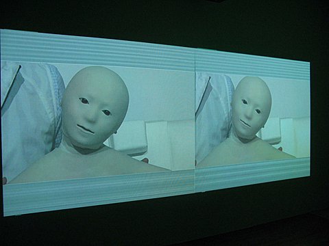 There is a large rectangular image projected onto the wall of a dark gallery room. The rectangular screen is broken in half and each half has an identical image. The image is of a masked AI mannequin like figure. The figure is staring at the camera with its head titled to the right. Behind the figure is another person standing of which we can only see their torso. The entire image is blue tinted and there are thick blue and white striped boarders on the top and bottom of the two side by side images.