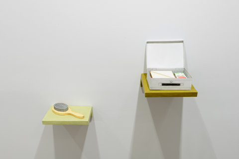 Installation view of two objects protruding from the white wall in the exhibition like shelves. The first shelf object is slightly lower than the one on the right. The first shelf is green and a yellow hairbrush sits atop it. On the right, the shelf is darker and a small white box made out of paper sits open with its lid leaning against the wall. 