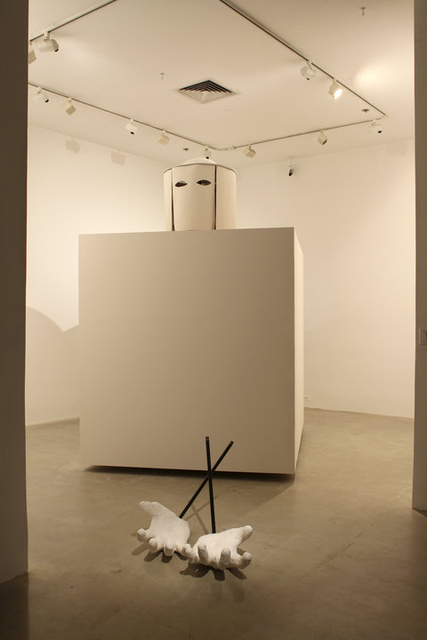 Installation image of a large white cube in the middle of one of the galleries. On top of the cube is a white cylinder with eye-holes cut out on the top. In front of the white cube are black poles crossed with two plaster cast hands, much larger than life, reaching out, palms up at the end of the poles. 