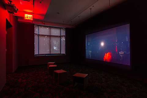 Three small benches sit in a dark room in front of a projection. A window on the wall is covered with a translucent film.