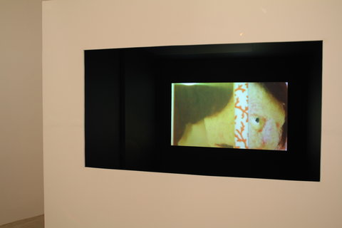 Close-up installation view of a black rectangle on the wall. In the middle, off center, is a video screen with an abstract green visual on view. 