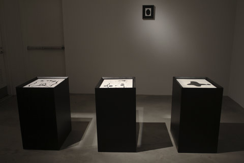 Installation view of a dark room with three pedestals in a row. Behind the pedestals, mounted on the wall is one black and white image. The image on the wall features a black frame, with a white piece of paper and a geometric black shape in the middle. The works on the pedestals are black and white drawings but are hard to make out from the cameras point of view.
