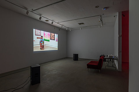 Two large speakers and a red couch sit in front of a grey table with a projector. A large projection on the wall shows a person reading from a book.
