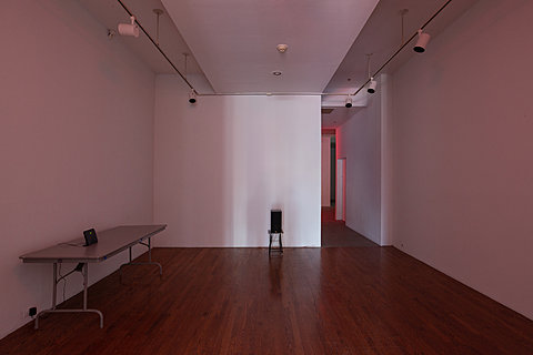 A speaker stands against a white wall; on the perpendicular wall a WiFi router sits on a grey table.