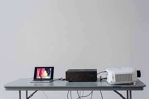 A close-up of a grey table holding a laptop, stereo reciever, and projector. The laptop screen shows a child's face, partially obscured.
