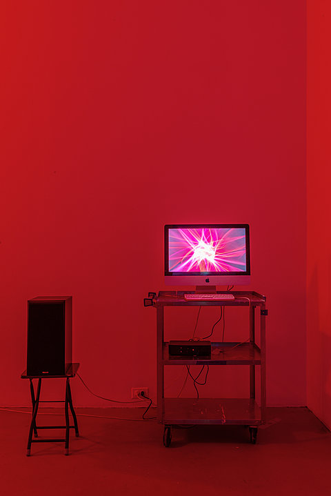In a room lit with red light, a computer displaying an animated screensaver sits on a desk next to a speaker.