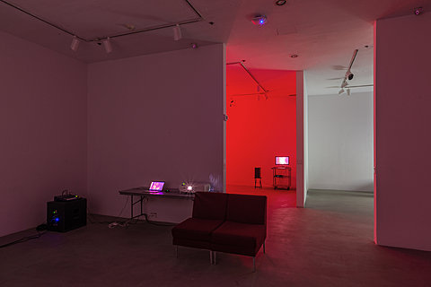 A room holding a mixing console, a table with a laptop and projector, and a red couch. In the background is an adjoining room filled with red light.