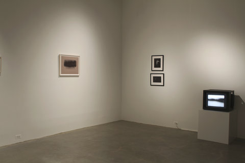 Installation view of two walls. On the right is a drawing hung on the wall. It is a tannish/peach piece of paper enframed in a white frame with glass. On the paper is a black shadow of charcoal in an organic, random rectangular shape. On the right wall are two stacked black frames. In each frame is a dark image that cannot be seen from the picture view. The image on top is vertical and the image on the bottom is horizontal. Next to the stacked frames, on the floor, is a pedestal with a small tv. On the tv screen is white background with a blurry black strike going through it. 
