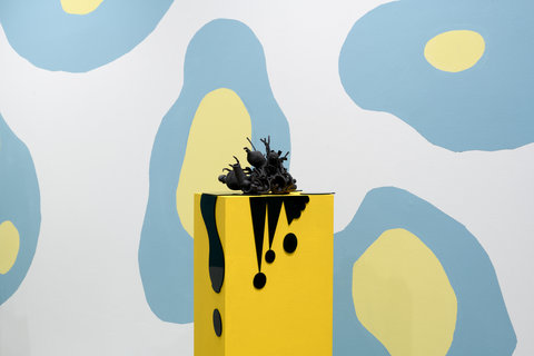 Installation view of a yellow pedestal in the exhibition. A black abstract sculpture sits atop the yellow pedestal. In the background, the white wall is painted with large organic shapes that are blue with yellow centers.  