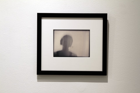 Close-up installation view of a black frame with a picture of a blurry shadow. The shadow resembles a figure from the shoulders up and is focused more towards the left side of the frame. 