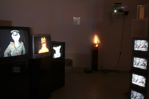 Installation view of the exhibition featuring a dark room with a projection on one wall. The projection is abstract, dark and blurry. There are several television monitors stacked on top of one another and placed throughout the room with various figures on the screens.  