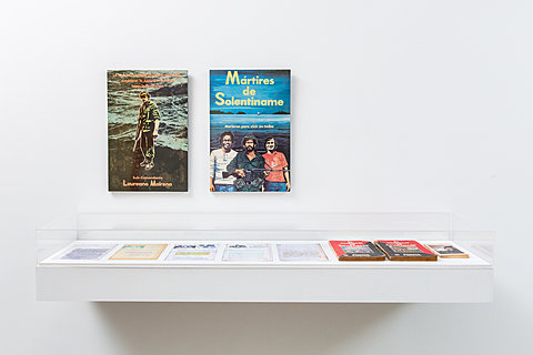 Two color posters hang above a wall-mounted glass-top case, holding several pamphlets and papers/