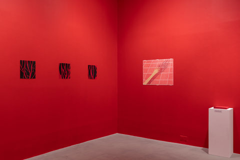 Installation view of the exhibition featuring two walls. On the left wall there is three drawings of red vein-like lines against a black background. The drawings are evenly spaced out. On the right side wall there is a drawing of light red squares separated by thin white lines. A figure's hand is outstretched over the squares. Next to the drawing on the ground is a white pedestal. Atop the pedestal is a pile of red cards. 