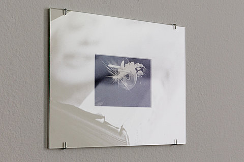 A tiny image mounted on a matted frame. A projection of a face on a book cover can be seen in the reflection of the frame.