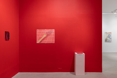 Installation view of the exhibition featuring two walls of a bright red room. On the left wall is one drawing that features red vein-like lines over a black background. On the middle wall, there is a drawing featuring light red squares at an angle separated by thin white lines. Over the squares is an outstretched figures hand. Next to this drawing is a white pedestal on the floor. Atop the pedestal sits a stack of red cards. There is a white room off to the left with another work in the exhibition in the background.  