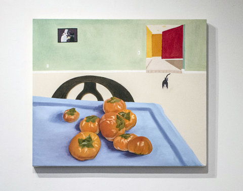 Image from a painting in the exhibition hanging on a white wall. The painting features an indoor scene with a corner of a blue table and a black chair tucked in. Orange fruit, similar to persimmon are scattered over the blue tablecloth. In the background, a green wall sits with a painting hung on it. The painting features two figures, a person and an animal from behind. A dog sits on the floor in the midground, turned away from the audience. A doorway is carved into the green wall revealing red and orange rooms that cascade back into the painting. 