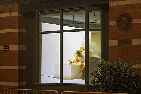 Shot from outside of a red brick building, a large projection can be seen on the wall through the window.