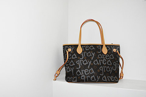 A fake Louis Vuitton monogrammed handbag. The handbag is brown with the words "grey area" written multiple times in grey ink across it.