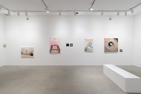 Installation view of several works hanging against a white wall in the exhibition space. In the gallery, on the right side of the room, a large white rectangular prism bench sits on the cement floor. 