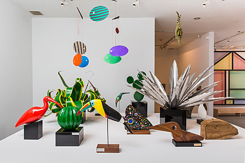 Several colorful sculptures, including sculptures of tropical birds and plants sit on a white surface. Two mobiles hang above the sculptures.