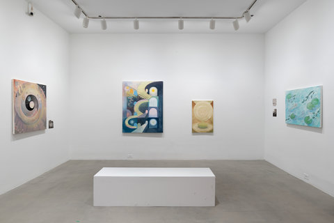 Installation view of four artworks in the exhibition hanging on three white walls. Two hang on the left wall, two in the middle and two on the right. In the center of the gallery space, a white rectangular prism bench sits in the middle of the floor. 