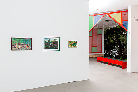 A wall with three paintings, in the background is a room with a red bench.