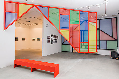 A gallery room with a red bench and a large, colorful geometric structure hanging from the ceiling. A further room with paintings can be seen in the background.