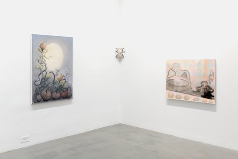 Installation shot of three works on two white walls. On the left wall, there is a work featuring fruitlike objects in the foreground, on the bottom, from their stems emerges long twisting dark stems that twist on the large green leaves and flower in the middle ground. The bulbous green leaves and stems have buds at the top. A large moon like orb sits in the background of the image, shining a light on the plants, and fruit-like objects in the foreground. The rest of the background is soft blends of blues. Where the two walls meet in the center of the image, is a small square artwork of two sketched figure's faces facing one another in profile view. They are drawn very simple, white with a black line. On the right wall, there is a pink canvas. On the bottom of the canvas, is a series of soft, blurred circles. Above, a figure sits at what seems to be a fountain. The figure is drawn very sketchily, just an outline, and lies it's head in the palm of its hand. Dark abstract lines and blobs are next to the figure on the right side of the painting. 