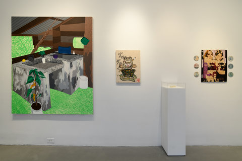 Installation view of one wall in the exhibition. A large abstract painting of what appears to be an outdoor garden scene of a shed. Bright green covers the background, with a suggestion of a stone table and wood shed. Next to the painting, a smaller painting on the left includes an painting of a frog wearing clothes, seated on a lily pad. The frogs arms are crossed in an anthropomorphic fashion. Next to the frog on the right, is a white pedestal, and next to that is a painting with three small circular discs on either side of it. 