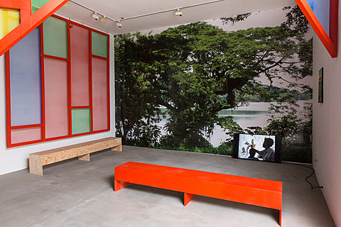 A small television monitor sits in the corner of a room in front of a red bench. The wall is covered with a print of a tree in front of a lake.