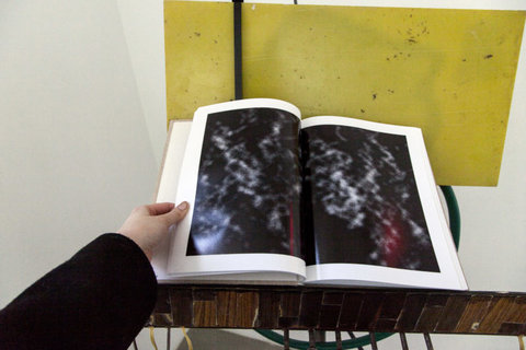 Image featuring an object in the exhibition. The image is framed as though the camera lens is the viewer looking down a a book in front of them. An arm wearing a black sleeve and white hand reaches and holds the left page of the book down. A yellow square sits behind the book against the wall. 