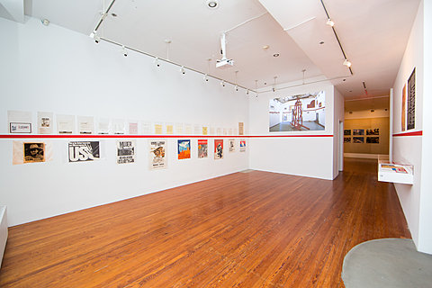 A strip of red paint runs horizontally across a gallery's walls. Images are displayed above and below the line. A projection of a similarly arranged room is on one wall.