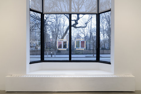 Installation view of the exhibition featuring the window in the gallery with four panels of glass and two artworks hung in the two center panels. In the left window pane, two abstract prints are taped to the glass and on the right side, two other prints are taped to the glass. 