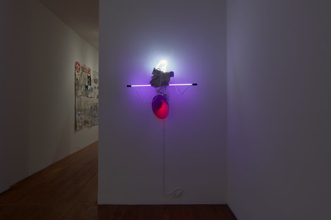 Installation view of a wall sculpture in the exhibition that features a neon, white, and red light mounted to the wall in an abstract conglomerate. 