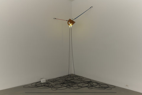 Installation image of a sculpture in the corner of the gallery. The sculpture stands on two wire thin legs and climbs up to meet a light-fixture looking object with two thin wirey poles protruding from the piece in opposite directions. The corner of the floor, where the walls meet is covered in wires weaving together around like snakes. 