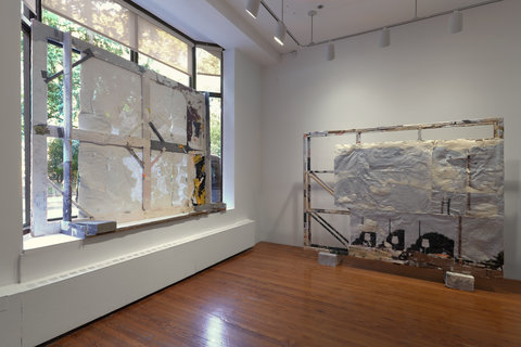 Installation image featuring a gallery room in the exhibition. A large white, canvas covers the windows on the left. On the right, another large unfinished looking canvas stands upright in front of the wall. 