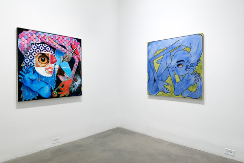 Installation view of two works in the exhibition. The left and right walls converge in the center of the image at the corner. On the left, the work is abstract and consists of several blue and pink colors with the main focal point being an eyeball. On the right wall, a blue fantastical figure is crammed into the edges of the canvas against a green background. 
