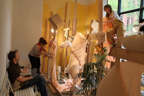 Installation view of the exhibition featuring a mish-mash of objects and artwork creations. The gallery room - turned construction studio features random wood planks and the center of the image focuses on a pale-skin like, peachy colored fabric stuffed with stuffing, slightly bursting at the seams. People work amongst the installation. 