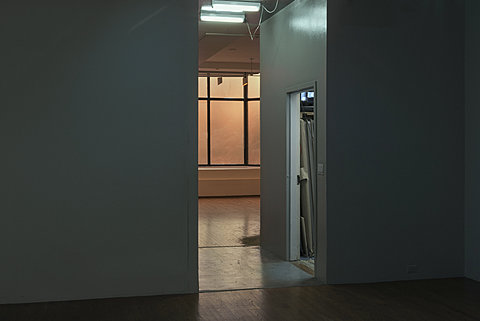 A room with warm light can be seen through a thin hallway from a darker room.