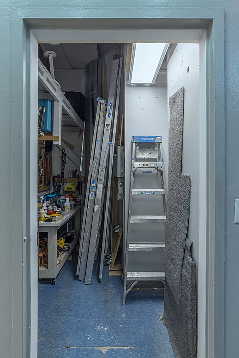 A flourescent-lit storage close holding ladders and carpet sections.