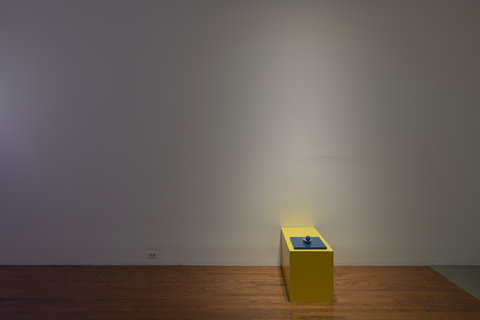 Installation view of a sculpture on the floor of the exhibition. The exhibition space is dim, almost no light shines except for a spotlight on the object that sits on teh woodo floor. The object consists of a box with a yellow top. Atop the box is an object that seems to be illegible form camera view. 