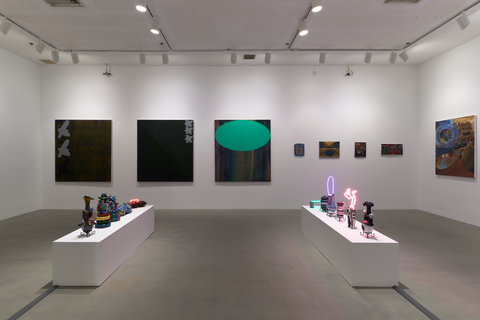 Installation view of the exhibition featuring three very large dark paintings on the left side of the back wall. The third dark, mostly black painting includes a teal oval on the top. On the right side of the back wall, four smaller dark paintings are hung against the white wall. Each work is illuminated by a white spotlight. In the foreground, two white, bench-sized pedestals sit on the ground with several sculptures lined on them. Some of the sculptures have neon light implements. 