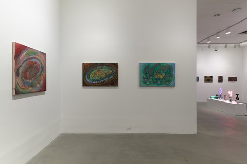 Installation image of three works hanging on white walls in the exhibition. On the left wall, a work is slightly illuminated by a white spotlight. On the right or middle wall, that is head on from the viewpoint of the camera angle, two paintings are with slightly illuminated spotlights on them. The background of the image leads to another room with more artworks. 