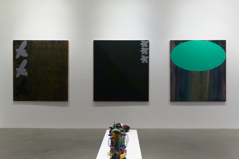 Installation view of the exhibition featuring three dark, mostly black paintings hanging on a white wall. While the first two are mostly black, the one on the right is black with a large teal oval, sitting horizontally painted on the top of the canvas. In the foreground, on a white bench pedestal, several sculptures sit in a row, but they are illegible from the camera view. 